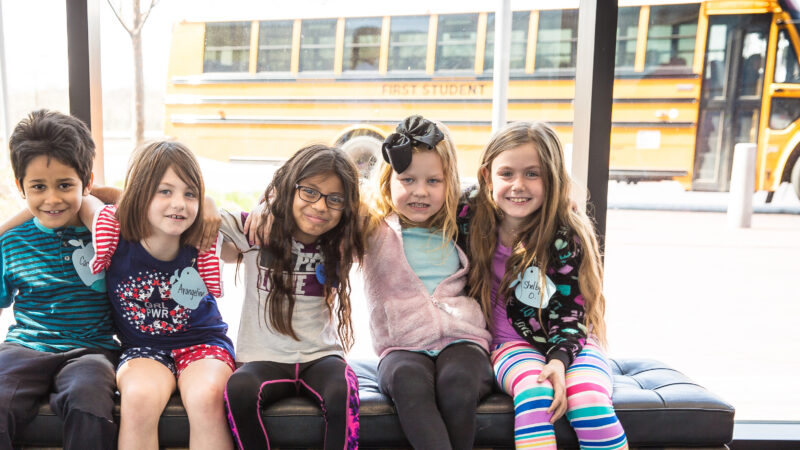 Five elementary-age students sitting on a black bench in the museum's Boeing Foyer with their arms around each other and smiles on their faces in front of the windows with a bus outside parked in the Lattner and Walker Family Plaza