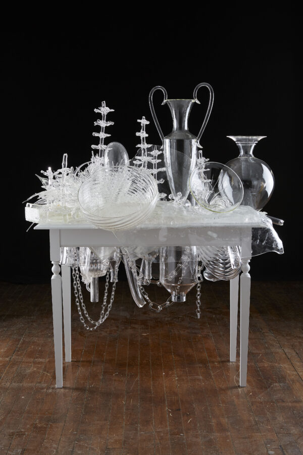 Sculpture of clear glass vases, vessels, bowls on a white table with more glass bowls, vessels, vases and chain link suspended underneath the table. The table is sitting against a solid black background with a worn, hardwood floor in red tones.