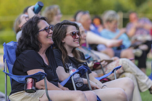 Photograph of two women sitting in lawn chairs in an outdoor setting. Both women are dark-haired, earing dark-colored clothing and sunglasses.