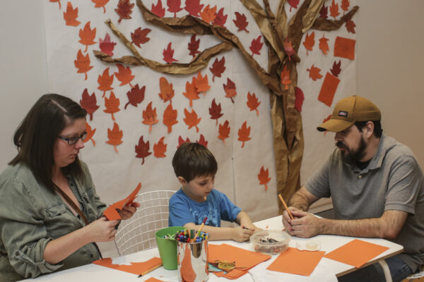 Photograph of two adults and a child sitting at a table with shades of light brown paper, markers, and scissors in front of them. An image of a tree with various leaves of browns and reds is on the wall behind them.