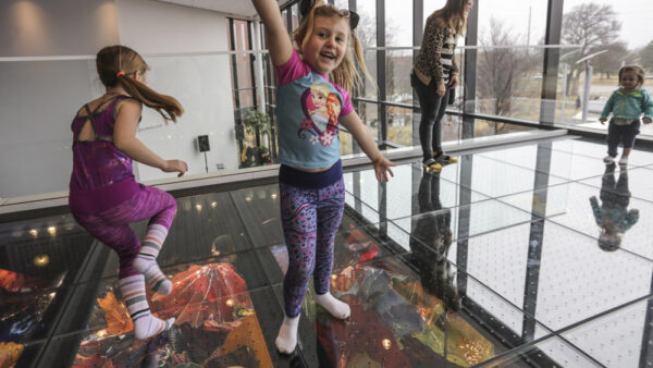 Photograph of a young girl with light brown hair in pigtails, standing with arms outstretched, wearing purple print leggings and a blue and pink shirt. She is standing in front of glass windows on a glass floor over bright clored, blown glass plates.