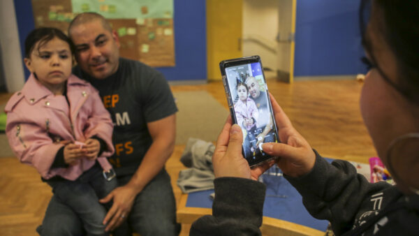 Photograph of man dressed in a black t-shirt and jeans, holding a young girl dressed in the pink jacket, while a woman dressed in a black jacket takes a photograph with a cell phone.