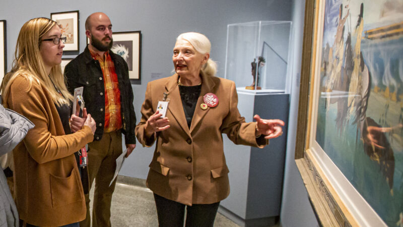 Photograph of people int the gallery. Woman at right has long blonde hair, dark-rimmed glasses and is wearing a tan coat. Man int he middle is bald with a dark beard, wearing an orange and yellow shirt under a dark jacket. Woman at right, standing in front of a painting, has white hair pulled back into a ponytail and is wearing a tan jacket over a black shirt. Her arm is outstretched with palm up toward the painting