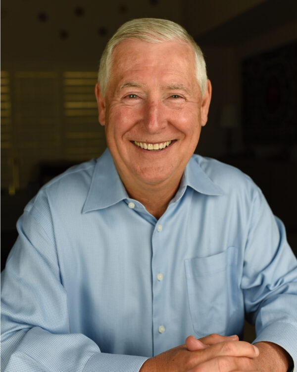 Smiling Man shown from approximately waist up, with arms in front and fingers interlinked. White hair, dressed in a blue button-up shirt