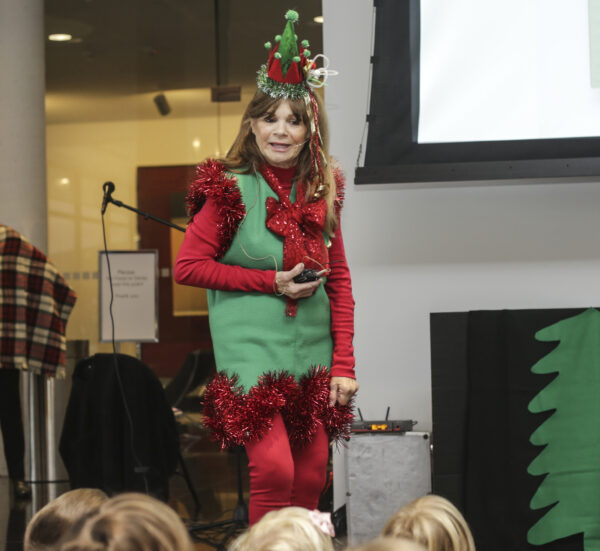 Woman stands speaking to several seated children while wearing a red and green crown, a green vest trimmed with red garland, a big red bow around her neck and red tights