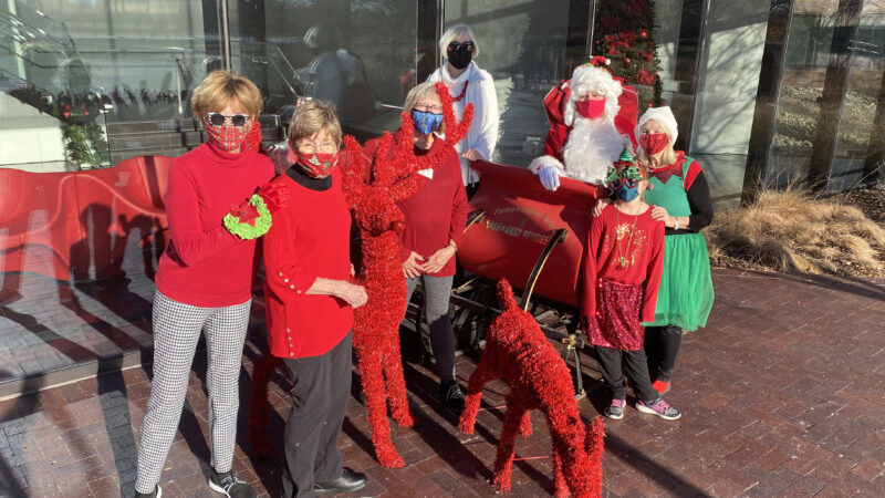 Group of people standing with Santa, Mrs. Claus and an elf around a red sleigh and red reindeer decorations