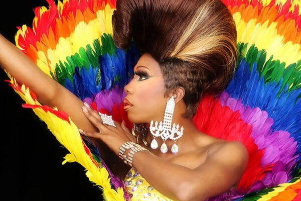 Profile of person with elaborate updo hairstyle against a colorful rainbow background. Arms are outstretched up to the left and she is wearing a long, glittery earring in an unside down T shape.