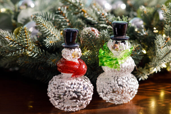 Glass snowmen on a background of a fir tree branch. Snowman on the left has a black hat, orange nose, and red scarf. Snowman on the right has a black hat, orange nose and green scarf.