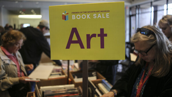 Yellow sign in the foreground with text Friends on Wichita Art Museum Book Sale, ART on a stick above boxes of books on a table. Two women in either side of the table looking at the boxes of books.