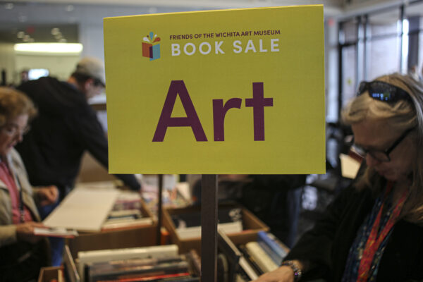 Yellow sign in the foreground with text Friends on Wichita Art Museum Book Sale, ART on a stick above boxes of books on a table. Two women in either side of the table looking at the boxes of books.