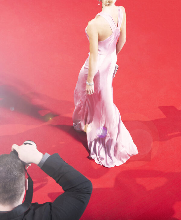 A white woman in a long pink dress with her back to the camera walks away from a white male photographer with short brown hair wearing a dark suit jacket and aiming a camera at the woman