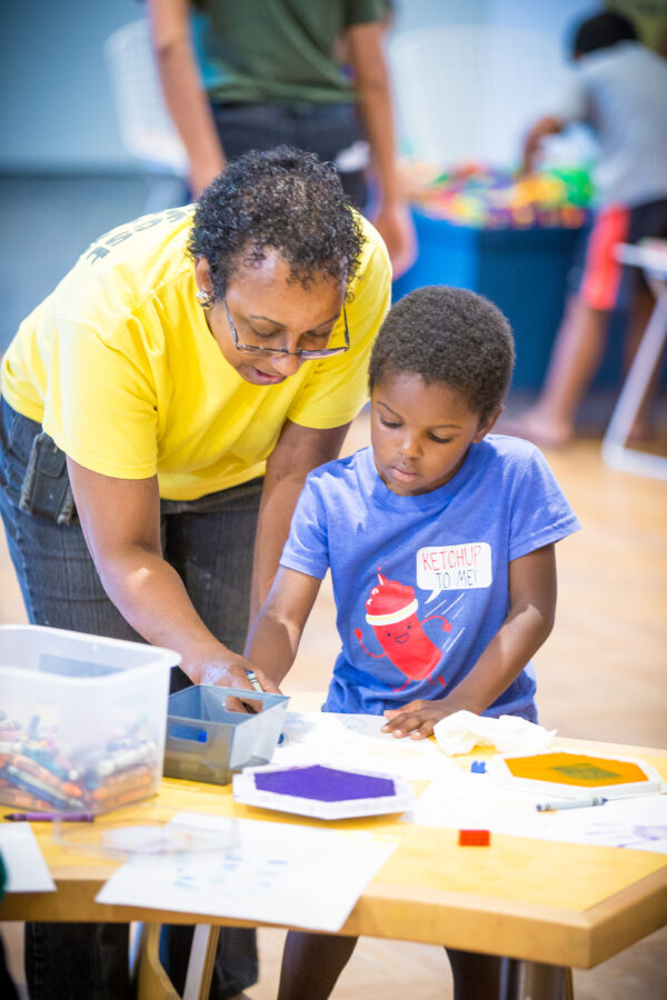 Adult in a yellow shirt, bending over the shoulder of a young boy working on an art project. Boy is dressed in a blue shirt with an image of a ketchup bottle and a balloon caption 