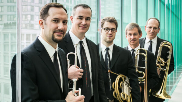 Five men dressed in dark suits, holding brass musical instruments, in front of a wall of windows with an cityscape int the background