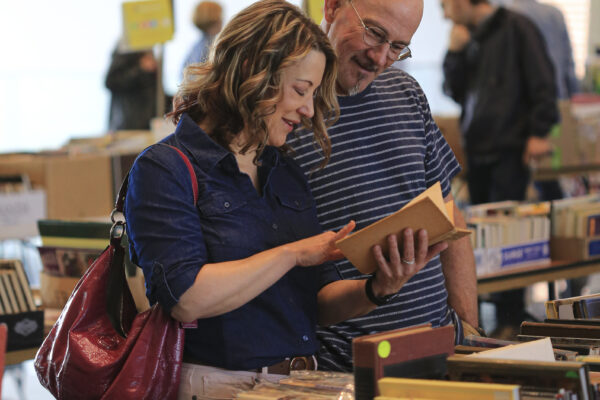 Photo of two people, a white woman with brown hair, blue shirt and raspberry purse, and a white man with glasses and blue/white striped shirt, looking through books spread out on tables