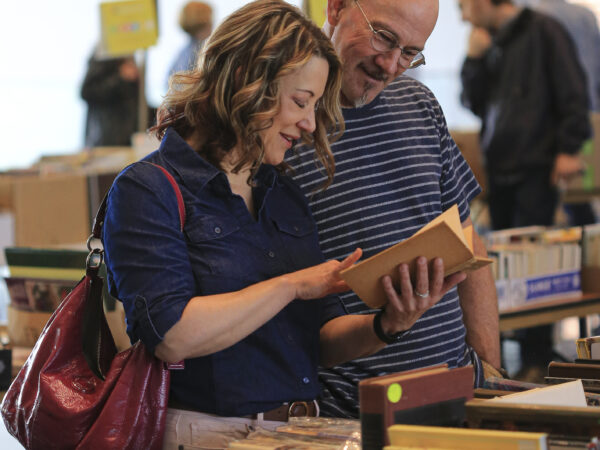Photo of two people, a white woman with brown hair, blue shirt and raspberry purse, and a white man with glasses and blue/white striped shirt, looking through books spread out on tables
