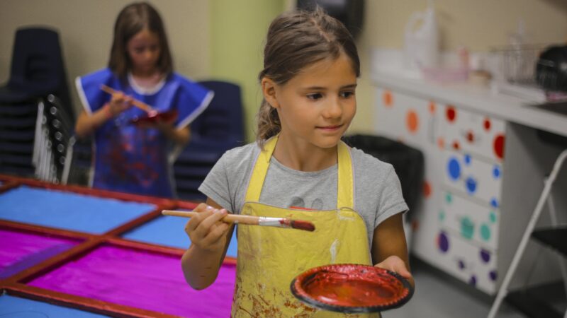 Young girl standing holding a paint brush in her left hand and a small plate with red paint in her right hand. Her brown hair is pulled back into a pony tail and she is weaing a gray shirt with a yellow, paint-stained apron.