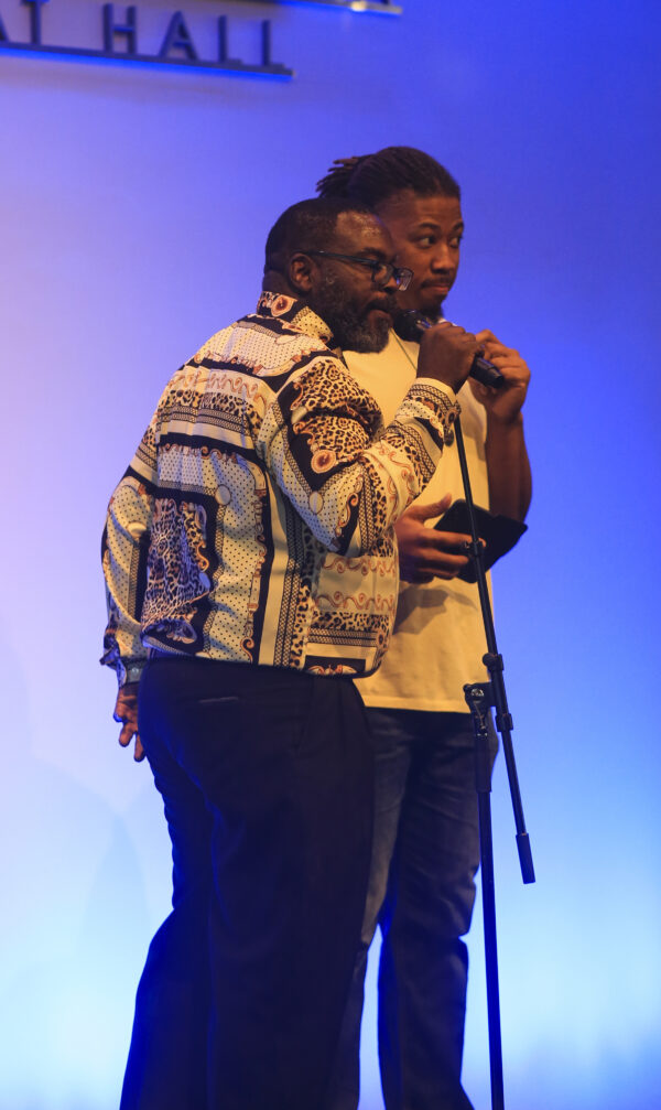 Two black men with microphones. Man on the left has close cropped hair and is wearing a cream colored shirt with wide stripes of a brown ethnic print. The man on the right has longer hair, pulled back and is wearing an ivory shirt.