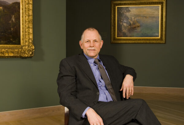 Man in a dark suit, blue shirt and tie, sitting in an art gallery