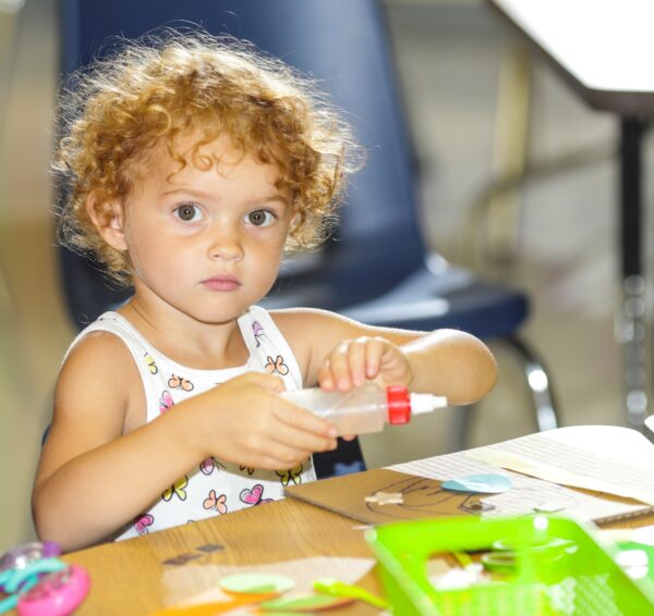 Young girl with curly red-blonde hair, dressed in a white tank top, sitting at a table with art supplies on the table in front of her and holding a bottle of glue with a red cap.