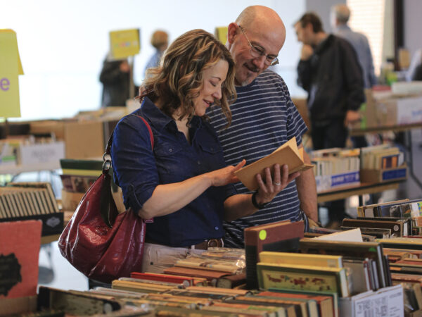 Couple standing in front of a tablecovered with books. Woman on the left holds a book in her hand and is pointing to a page inside the book. She is dressed in a blue button blouse and has a red leather bag over her right shoulder. The man looks over her left shoulder at the book. He is balding, wears glasses and and a blue and white striped shirt.