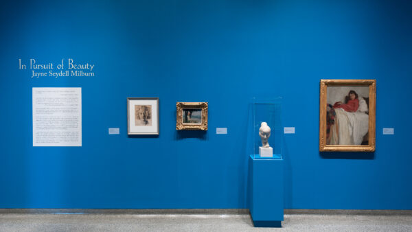 Blue gallery wall with one large, white didactic panel, two gold-framed paintings, one framed drawing, and one white marble sculpture of a woman's head under a vitrine on a blue pedestal