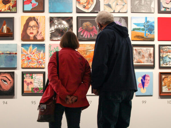 A man on the right in a dark blue jacket and jeans stands next to a shorter woman on the left wearing a red jacket, jeans and carrying a purse, as they both stand in front of 38 12x12 paintings on the wall