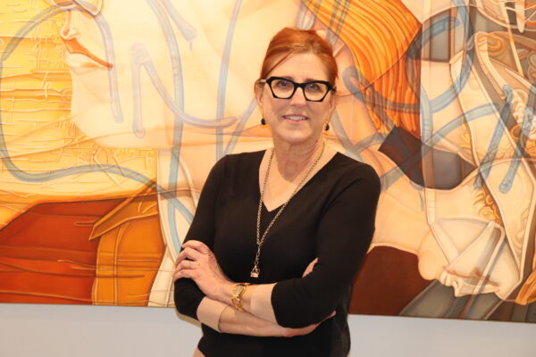 Red-haired woman with dark-framed glasses, dressed in a black v-neck with a long gold necklace, standing in front of an abstract art in yellow and orange tones