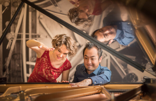 Woman and man framed by the lifted cover of a grand piano. Both are looking down, woman has an arm raised. She is dressed in a red sleeveless top and has light brown curly hair. The man, on the right, has one hand on top of the piano and is wearing a long-sleeved blue shirt