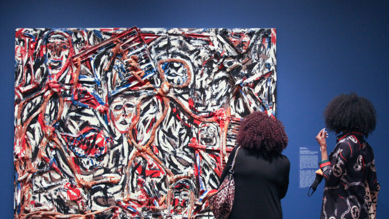 Two black women, one in a black dress carrying a leopard purse and the other searing a black dress with red lines and white, circular shapes, stand in front of a rectangular abstract artwork painted with the colors white, red, black, blue and dark orange, hung against a blue wall