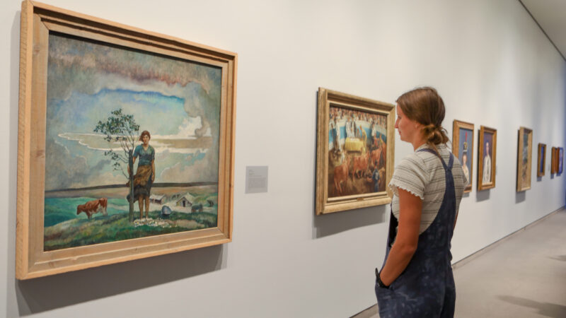 White woman with dark blonde hair, striped shirt and jeans overalls looking at paintings on the wall