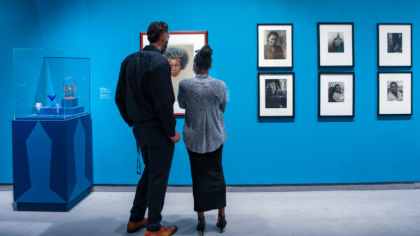 Man and woman standing in gallery in front on a painting of an African American man. On their left is a pedestal with an enclosed top an several pieces of glass. On their right is a collection of six photographs. Gallery walls are painted a bright blue. and six photographs
