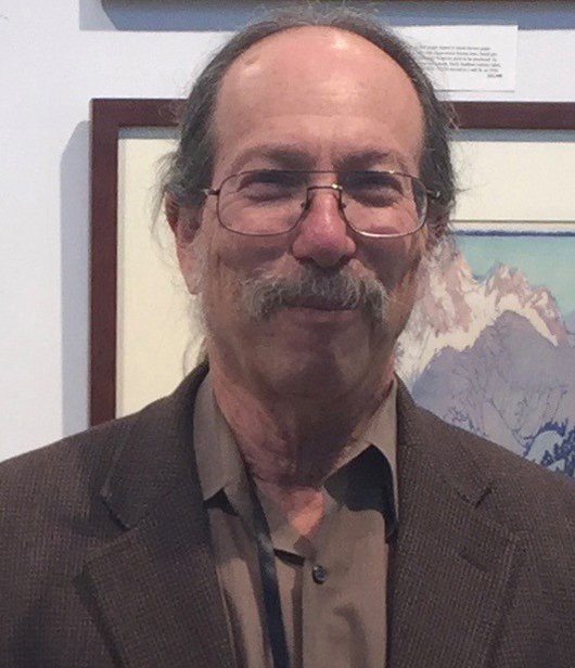 Head and shoulders of a white man, receeding hairline with brown hair, wearing rounded frame wire-rimmed glasses and with a brown and gray mustache and closely trimmed beard. Dressed in a brown jacked and open-necked brown shirt. Background is the corner of a framed artwork with blue and pink mountains.