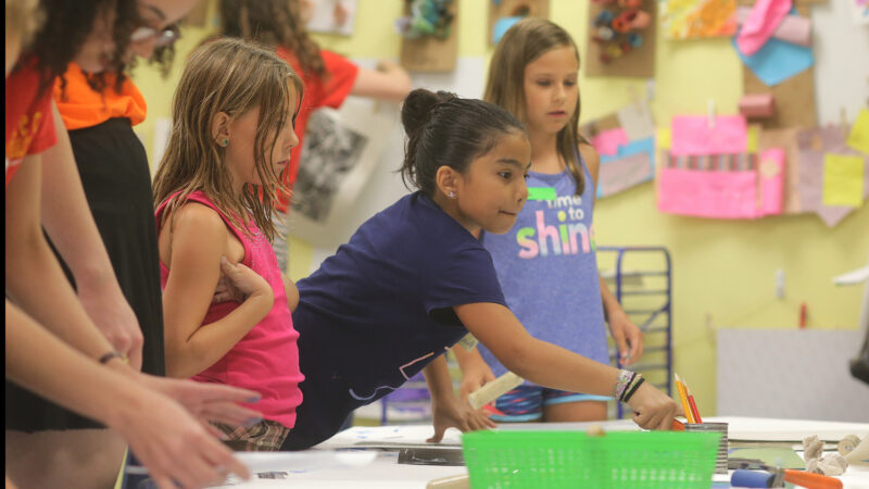 Three girls in the foreground wearing t-shirts in pink, purple and lavendar listen to artmaking instructions in front of a wall filled with student artwork
