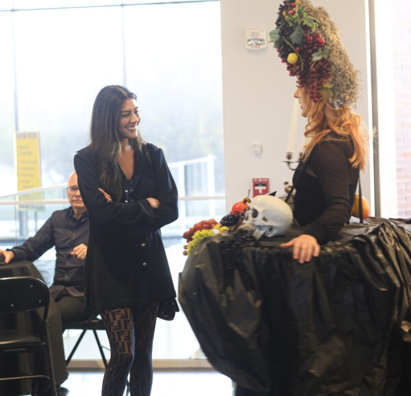 Two women stand talking to one another, one dressed in an all-black ensemble and the other wearing a table around her waise and a pile of fruit in her red hair