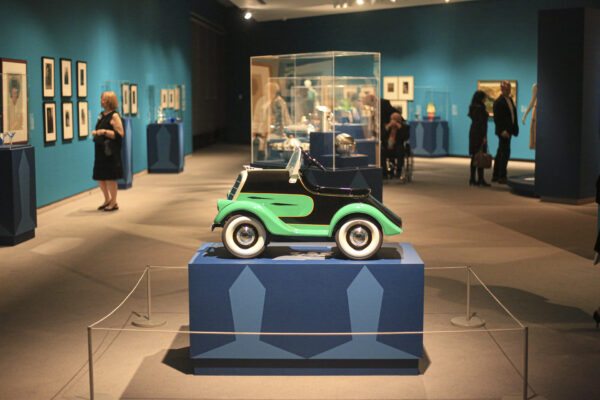 Gallery image with a green and black pedal car on a blue pedestal in the foreground, with people in the gallery in the background