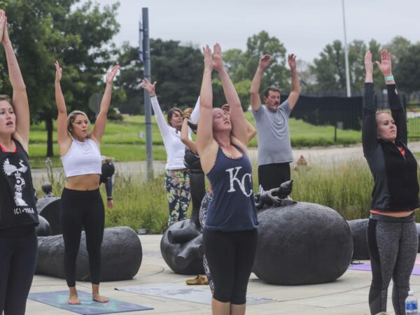 Group of people in standing in a yoga post with arms over their heads in a garden setting, standing on a concrete pad with pieces of a black sculpture.