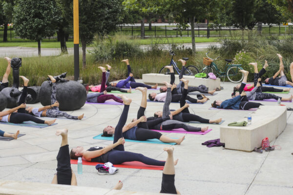 Exterior photo of the Jayne Milburn Sculpture Plaza where nearly two-dozen people are laying on the concrete plaza on colorful mats with their left legs stretched up in the air in a yoga pose