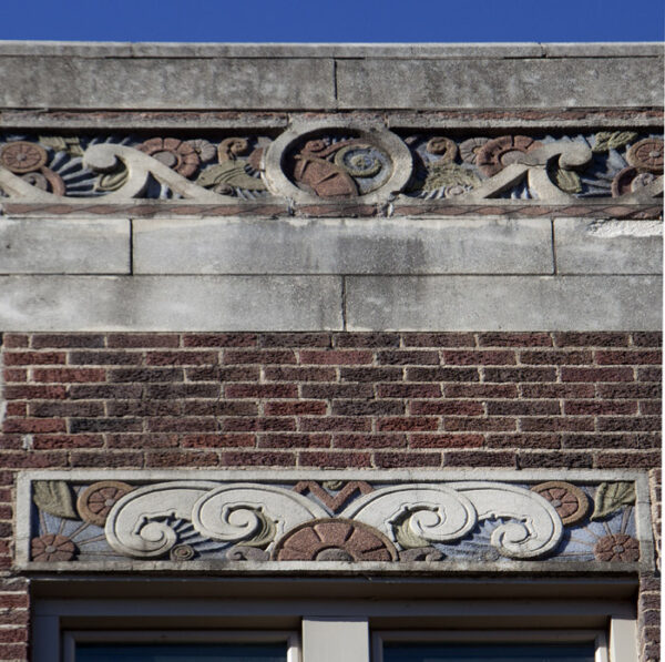 Buildling facade of red brick with a rectangle relief sculpture suggestive of white clouds and a floral motif in red and green leaves in the corners. Above is a similar relief with white swirls, red flowers and green leaves