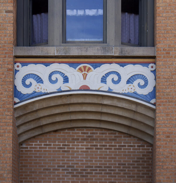 Relief sculpture in Art Deco style depicting clouds in white on a blue background around a rising sun in geometic patterns of orange and yellow, over concrete arches on a red brick building. Windows are the top of the imagender the scultou