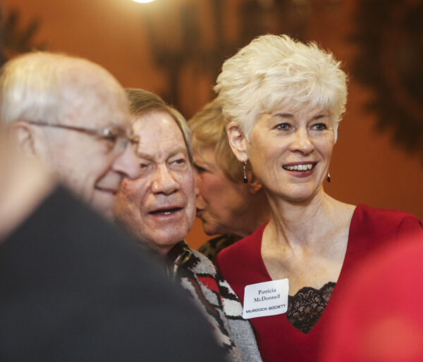 Group of people, from left, man in profile with fringe of white hair and brown-framed glasses, center, man with sand colored hair, dressed in brightly colored shirt, on left, woman with short white hair and dressed in red with name tag Patricia McDonnell