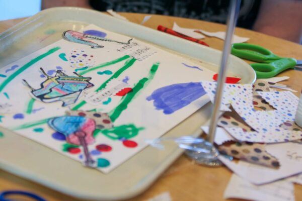Colorful abstract drawing with green lines, purple and blue shapes, on an off-white tray with pencils, other art supplies, and other drawings on the tabletop around it.