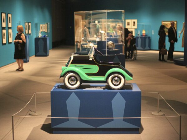 Gallery picture with a 1930s green and black pedal car on a two-tone blue pedestal