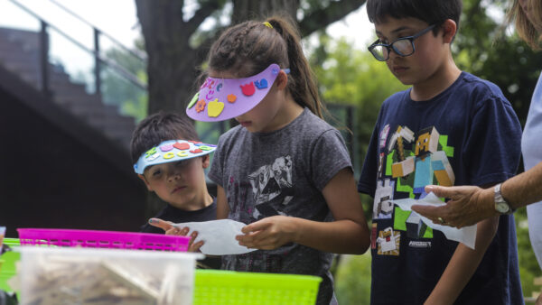 Three children outdoors doing an art project. At left, child is wearing a white sunvisor decorated with flowers. Girl in the center is looking down at a white peice of paper in her hands. Her face is mostly obscured by a purple sun visor with yellow, orange, red, and blue flowers. Her dark hair is pulled back into a pony tail. She is dressed in a short-sleeved dark gray shirt. At right is a dark-haired boy with blue-framed glasses and dressed in a navy shirt with a graphic design in greens and blues.