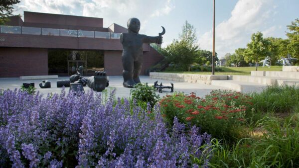 Garden photo with purple flowers in the lower left corner and red flowers on the right size. In the mddle of the image is Tom Otterness sculpture 