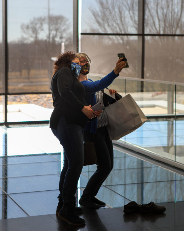 Two white women standing on the Chihuly Bridge, one with curly brown hair, long-sleeve black shirt, jeans and black boots holding a grey-case smartphone, the other with short blonde hair, glasses, long-sleeve blue shirt, dark pants and dark shoes carrying a white paper Museum Store shopping bag and taking a selfie of the two with her smartphone
