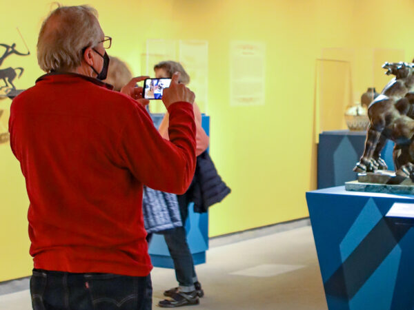 Photo of a white man with grey hair, glasses, a mask, red sweater and dark pants uses his smartphone in the galleries to photograph a bronze horse sculpture on a blue pedestal with three museum visitors in the background
