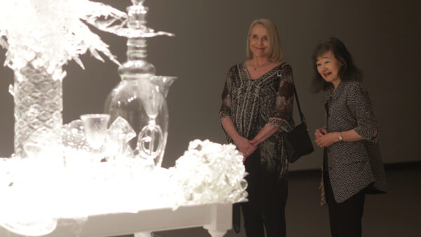 Gallery image on the right is a white table covered with clear glass vases, bowls, flower-like objects.., Left half of the image is dark with shadowed figure of two women