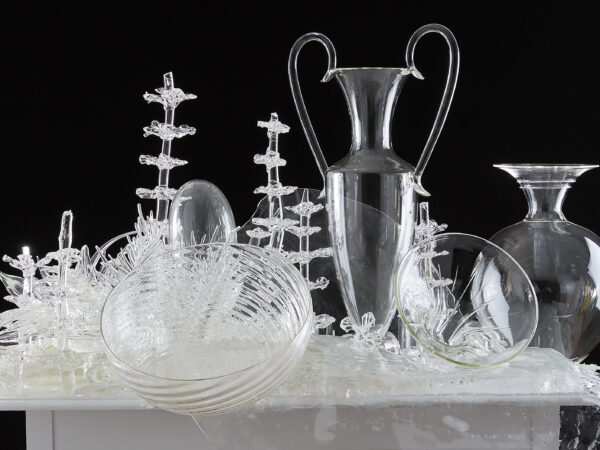 White tabletop covered in artworks of clear glass, including two vases, two bowls, and plant-looking forms