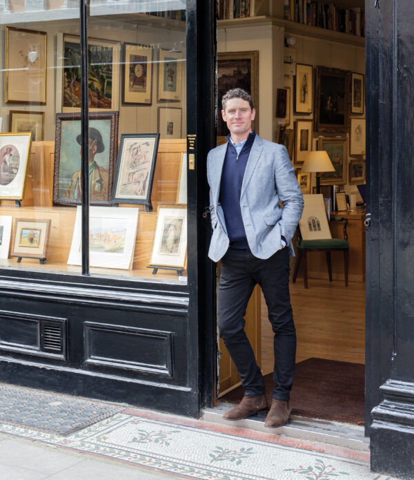 Man dressed in a light blue jacket and dark slacks, standing in the doorway of a shop. Varous artwork is vivible in the shop window to his raight.