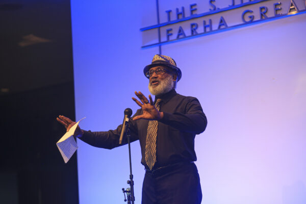A black man with a beard and glasses, wearing a black and tan hat, black long-sleeve shirt, black pants, and black and tan print tie, stands in front of a microphone with his arms outstretched, holding a piece of paper in his right hand